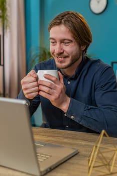 Young businessman working on laptop computer drinking cup of warm hot coffee sits at table workplace home office room in the morning. Male freelancer guy enjoying relaxing, taking a break. Vertical