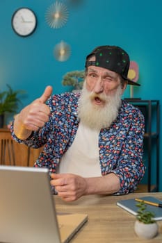 Like. Smiling stylish senior old man looking approvingly at camera showing thumbs up sign positive something good great news positive feedback. Happy elderly grandpa with laptop at home office desk.