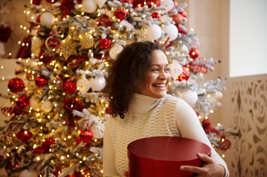 A joyful woman holding a red gift box, smiling in front of a beautifully decorated Christmas tree with lights and ornaments. The image captures the festive atmosphere and holiday cheer.