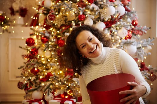 A happy woman holding a red gift box during the Christmas season, with a beautifully decorated tree and festive ornaments in the background. Celebrating the holiday spirit and joy.