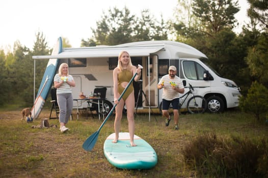 the family is resting next to their mobile home. My daughter is standing with a paddle on a sup board, and her parents pour water on her.