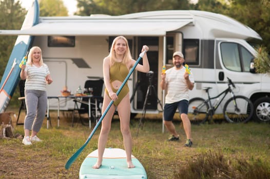 the family is resting next to their mobile home. My daughter is standing with a paddle on a sup board, and her parents pour water on her.