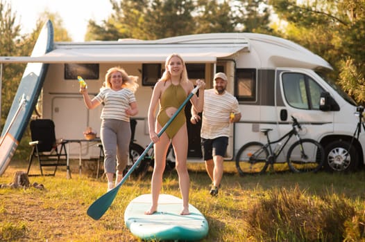 the family is resting next to their mobile home. My daughter is standing with a paddle on a sup board, and her parents pour water on her.