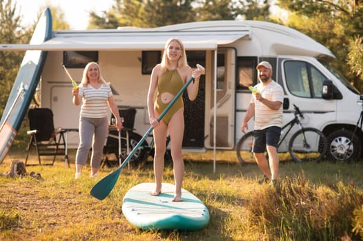 the family is resting next to their mobile home. My daughter is standing with a paddle on a sup board, and her parents pour water on her.