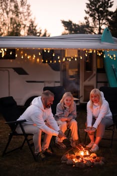 A family cooks sausages on a bonfire near their motorhome in the woods.