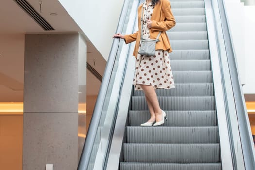 Unrecognizable young woman in a beige dress with large polka dots goes down an escalator in a shopping center, blurred background
