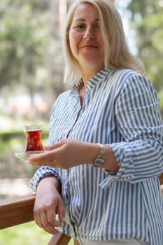 young blonde Caucasian woman drinks tea from Turkish Armudu cup on the balcony and takes bite of orange cake, delicious dessert after lunch, traditional oriental sweet on summer vacation