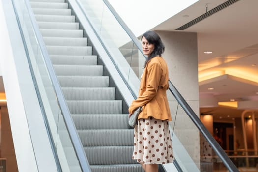 Confident young brunette goes up the escalator to the floor above in a shopping center, looks at the camera, blurred background, High quality photo
