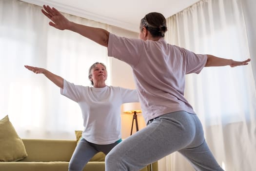 Senior woman exercising at home together with daughter. Active mature woman doing stretching exercise in living room at home.