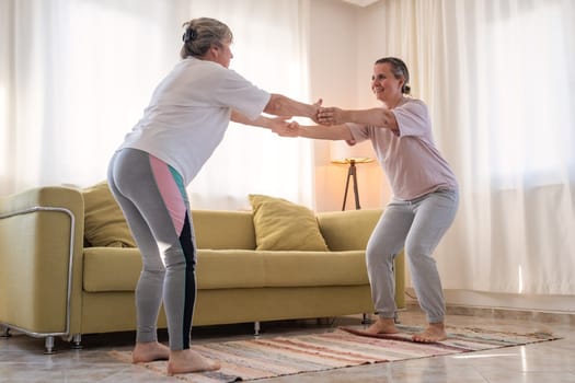 Senior woman exercising at home together with daughter. Active mature woman doing stretching exercise in living room at home.