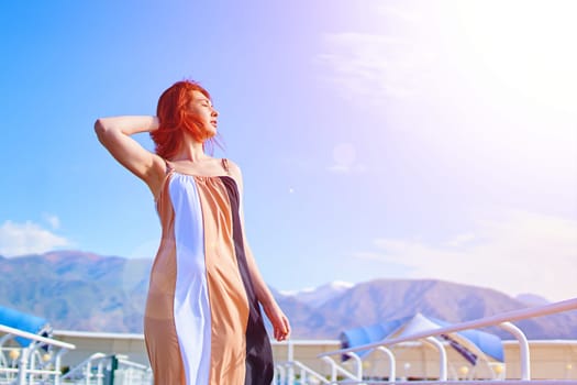 Woman in a dress stands on a pier on the beach by the ocean in a hotel against the background of mountains basking in the sun