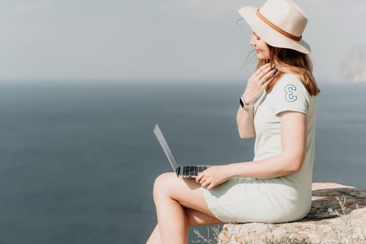 A woman in a sundress and hat sits on a cliff overlooking the ocean, using her laptop.