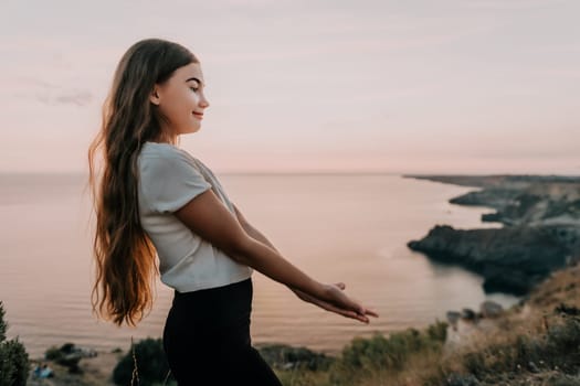 A young girl stands on a cliff overlooking the ocean at sunset, her long brown hair flowing in the breeze.