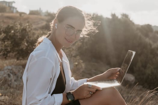Digital nomad, woman in the hat, a business woman with a laptop sits on the rocks by the sea during sunset, makes a business transaction online from a distance. Freelance, remote work on vacation.