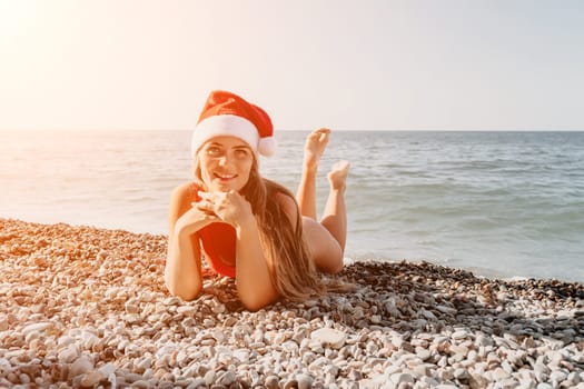 A young woman in a red swimsuit and Santa hat lies on a pebble beach, looking off to the side.