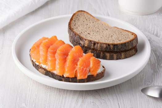 Rye sandwich with salmon and cream cheese on white wooden table.