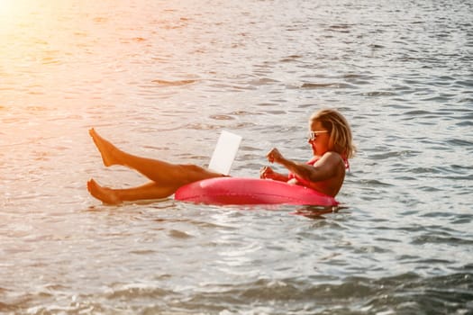 Woman works on laptop in sea. Freelancer, young blond woman in sunglases floating on an inflatable big pink donut with a laptop in the sea at sunset. Freelance, travel and holidays concept