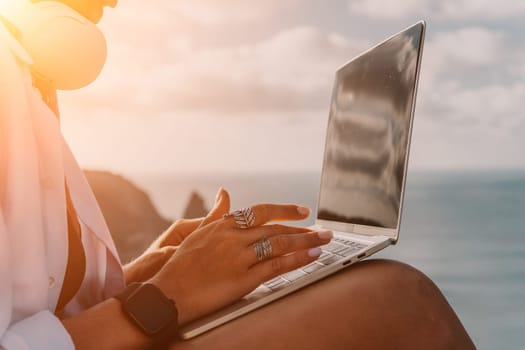 Digital nomad, woman in the hat, a business woman with a laptop sits on the rocks by the sea during sunset, makes a business transaction online from a distance. Freelance, remote work on vacation.