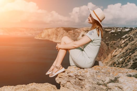 A woman in a hat and dress sits on a cliff overlooking the ocean, bathed in the warm glow of the setting sun.