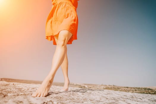 A woman wearing a red dress walks across a rocky clifftop with a bright blue sky in the background.