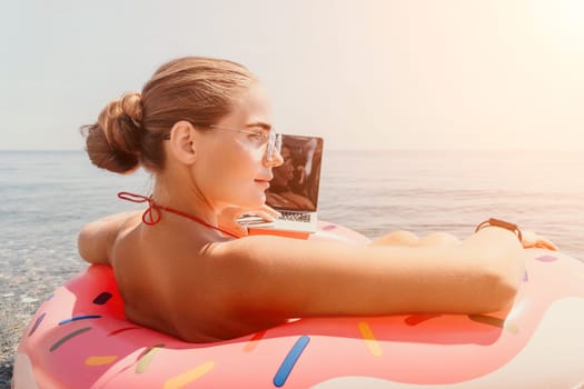 Woman freelancer works on laptop swimming in sea on pink inflatable ring. Pretty lady typing on computer while floating in the sea on inflatable donut at sunset. Freelance, remote work on vacation