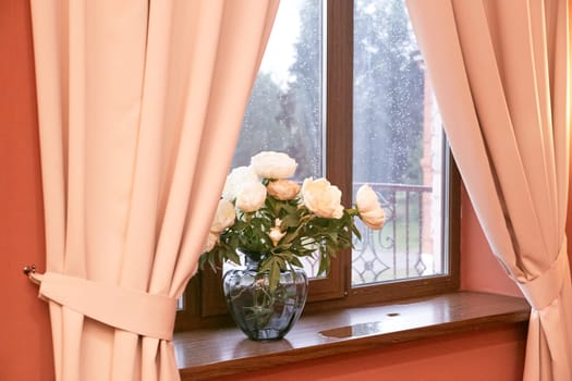 Cream peonies in a glass vase on the window. Flowers at home. Cosiness.