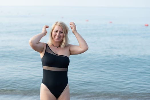happy young woman in a fashionable black swimsuit stands on a sandy beach by the sea on a beautiful sunny day, straightening her wind-tumbled hair