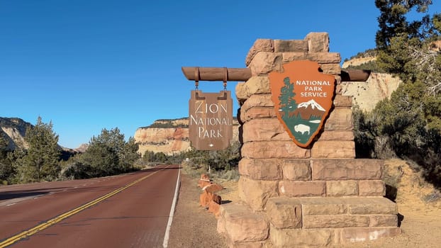 East Zion, Utah, USA – 02.28.2024: Scenic view of the Zion National Park east entrance sign on a bright sunny day. Lush green trees and shrubs line a red paved road leading into the park.