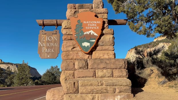 East Zion, Utah, USA – 02.28.2024: Close up of the Zion National Park east entrance sign. It is a beautiful day with blue sky. The sign is made of stone and wood. It is located on the side of a road.