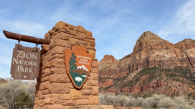 Springdale, Utah, USA - 02.27.2024: The main entrance sign for Zion National Park in Utah. The red rock sign is set against a beautiful blue sky, with the majestic Watchman mountain in the background.