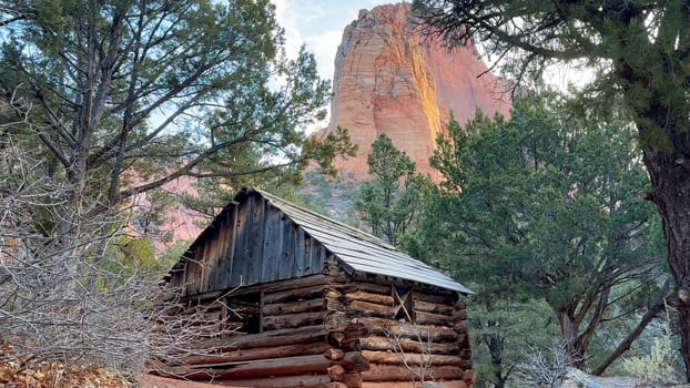 Weathered and rustic log cabin known as the Larson Cabin nestled among the trees of Zion National Park with a towering red rock mountain in the background - Taylor Creek, Kolob Canyons, Utah, USA