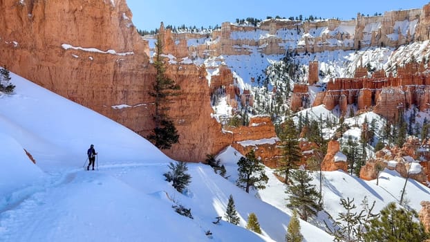 A female hiker in the distance on a sunny day hiking on a snow covered trail in Bryce Canyon National Park. The trail is surrounded by distinctive red rock hoodoos - Utah, USA