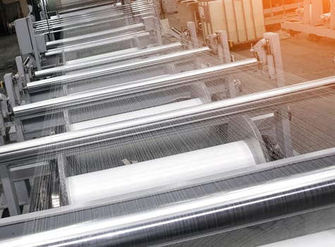 An industrial knitting machine in a textile factory with spools of white thread on a production line. Weaving fabric from yarn in a clean, well-lit manufacturing environment.