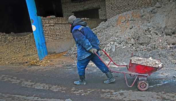 A worker in blue overalls and boots pushes a wheelbarrow full of debris on a construction site. The sun casts a warm glow on the scene, creating a sense of hard work and determination.