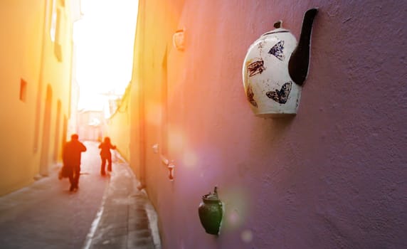 A white teapot with butterfly designs is attached to a pink wall in a narrow alleyway. Two figures walk away from the camera in the background, as the sun shines brightly at the end of the street.
