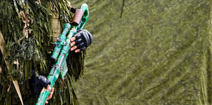 A soldier dressed in camouflage holds a rifle, standing near a green tent.