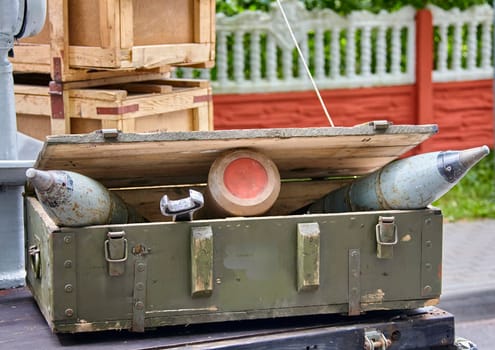 A military munitions crate is open with several large munitions inside, and the lid propped open. A fence and brick wall are in the background.