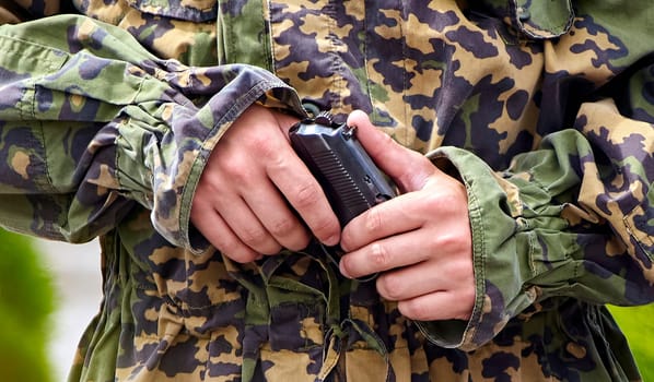 A close-up shot of a soldier's hand gripping a handgun. The soldier is wearing a camouflage uniform and is standing in a grassy area. The focus is on the hand and the gun, and the background is blurred.
