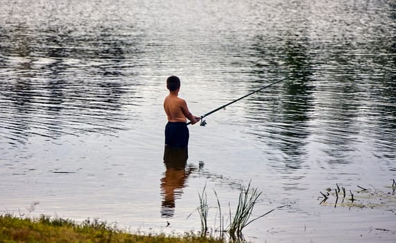A young boy stands in a lake, holding a fishing rod and casting his line into the water. The sun reflects off the rippling surface.