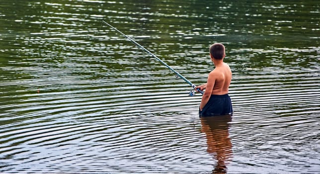 A young boy stands in a lake, holding a fishing rod and casting his line into the water. The sun reflects off the rippling surface.