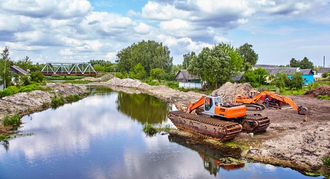A bright orange excavator sits beside a calm river, its tracks resting on the sandy bank. The river winds its way under a metal bridge in the distance, passing through a lush green landscape.
