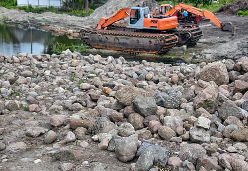An orange and gray amphibious excavator works on the edge of a pond, placing large rocks to stabilize the shoreline. The excavator's tracks are partially submerged in the water.