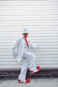 An elderly man in a white suit, huge boots and a clown nose walks funny