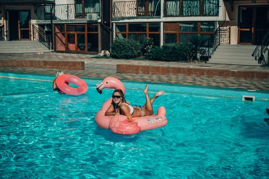 A woman is floating in a pink flamingo float in a pool. The pool is surrounded by a brick wall and has a few other people in the background
