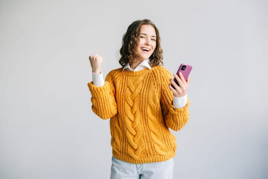 A joyful young woman student or professional, celebrating a victory on her smartphone. Standing against a white background, she expresses happiness and excitement upon receiving great news or an offer