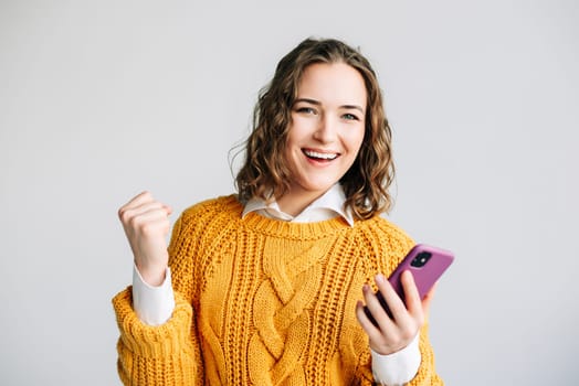 A joyful young woman celebrating good news on her smartphone. Standing against a white background, she radiates happiness and excitement, capturing the moment of receiving a positive message.