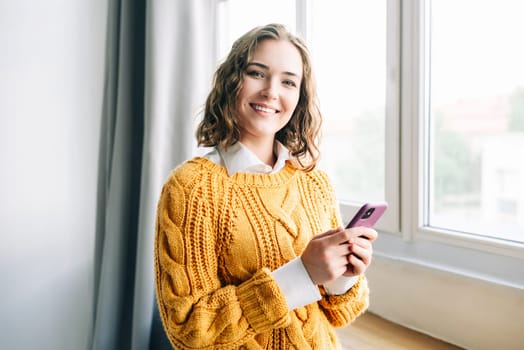 Smiling young woman using her smartphone at home, enjoying online shopping, messaging, and browsing. Capturing the essence of modern lifestyle and technology, she stands in a cozy indoor setting.