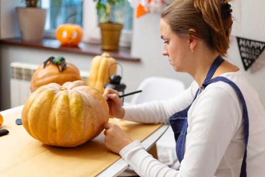 Woman carves a large pumpkin at a table, preparing for Halloween festivities, with other decorated pumpkins in the background.