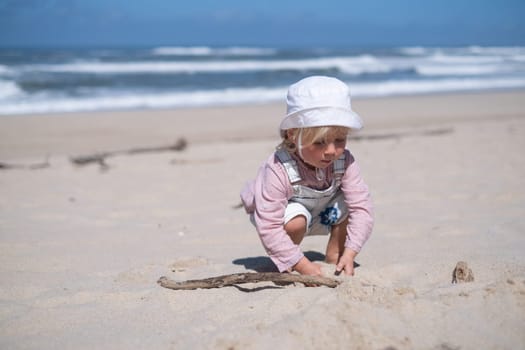 Little girl is sitting on sand and playing at sea beach in warm sunny summer day