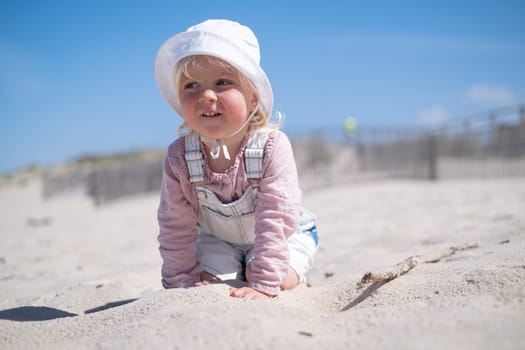 Little girl is sitting on sand and playing at sea beach in warm sunny summer day